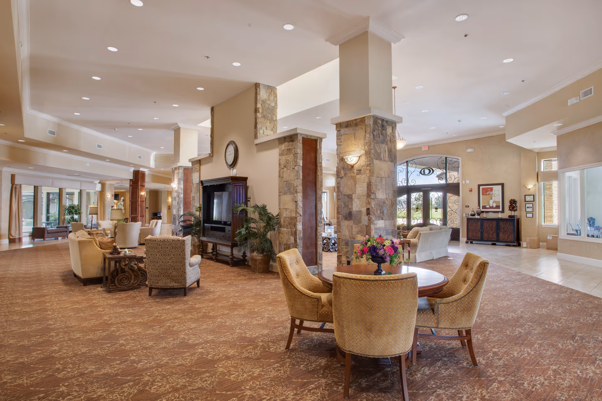 Spacious and well-lit senior living facility common area with comfortable seating arrangements including armchairs and sofas. The room features stone pillars, a large clock above a fireplace, decorative plants, a round table with four chairs and a floral centerpiece, and large windows and glass doors allowing natural light to enter.