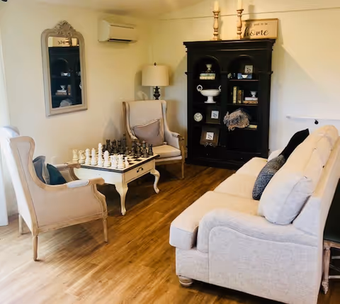 A cozy living room with a beige sofa and two armchairs surrounding a coffee table with a chess set. There is a black cabinet with decorative items and a lamp on a side table. A mirror hangs on the wall, and the floor is wooden.