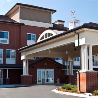 Entrance of a senior living facility with a covered driveway supported by white columns and a brick base. The building has multiple windows and a cupola with a weather vane on top under a clear blue sky.
