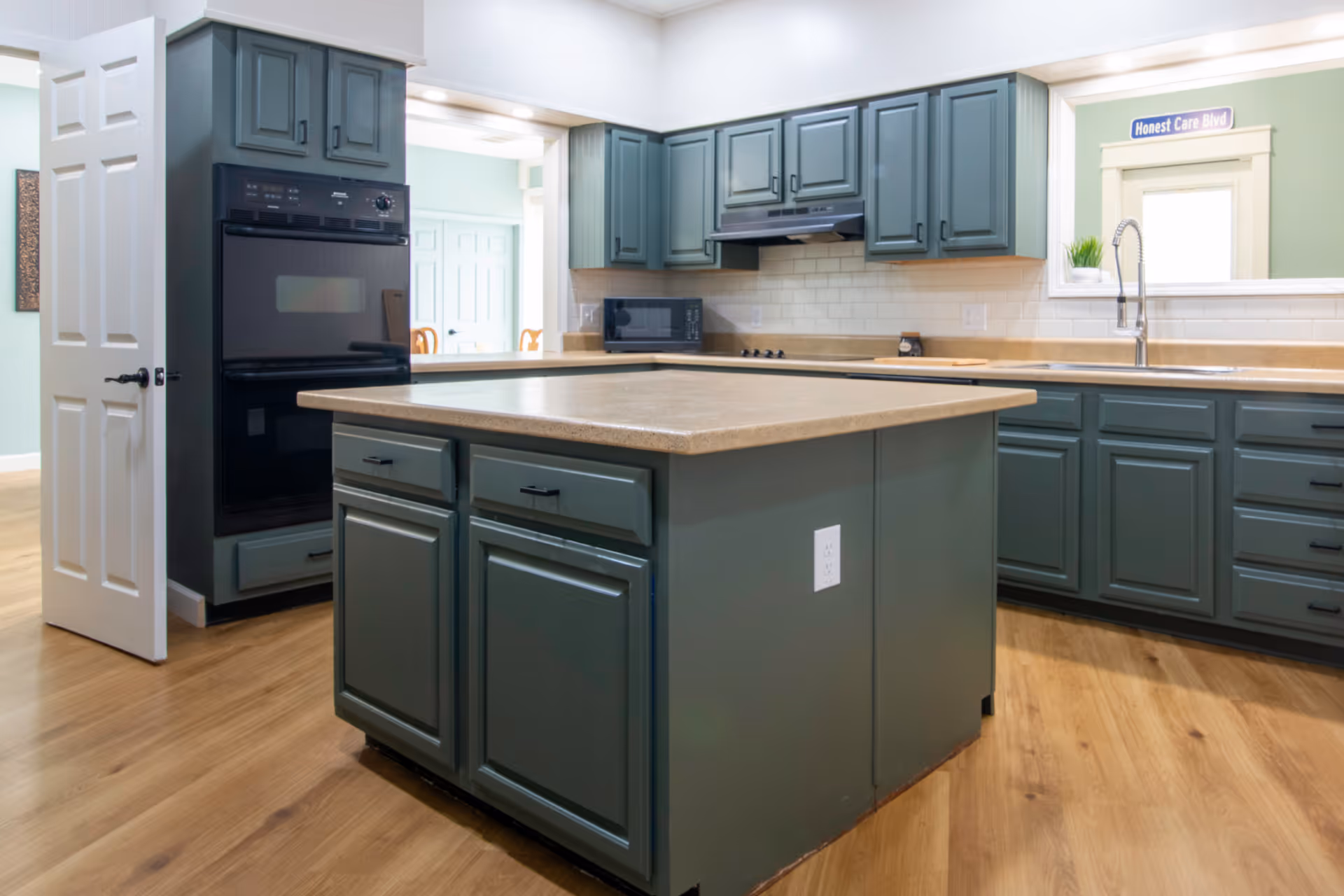 Spacious kitchen with teal-green cabinetry, a central island, double oven, and sink under a pass-through window.