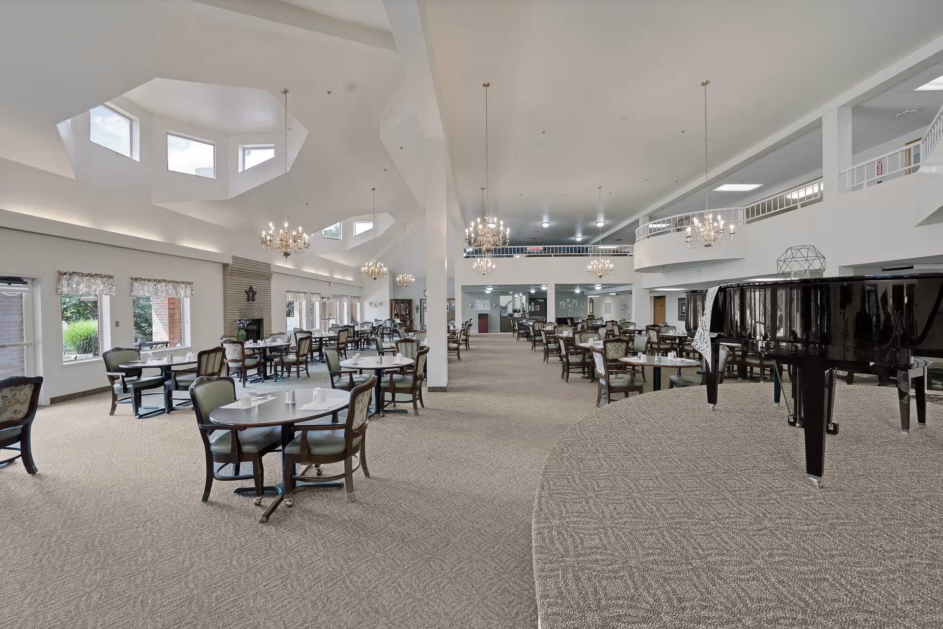 Spacious dining hall with round tables and chairs, chandeliers, large skylights, and a black grand piano on a raised platform.