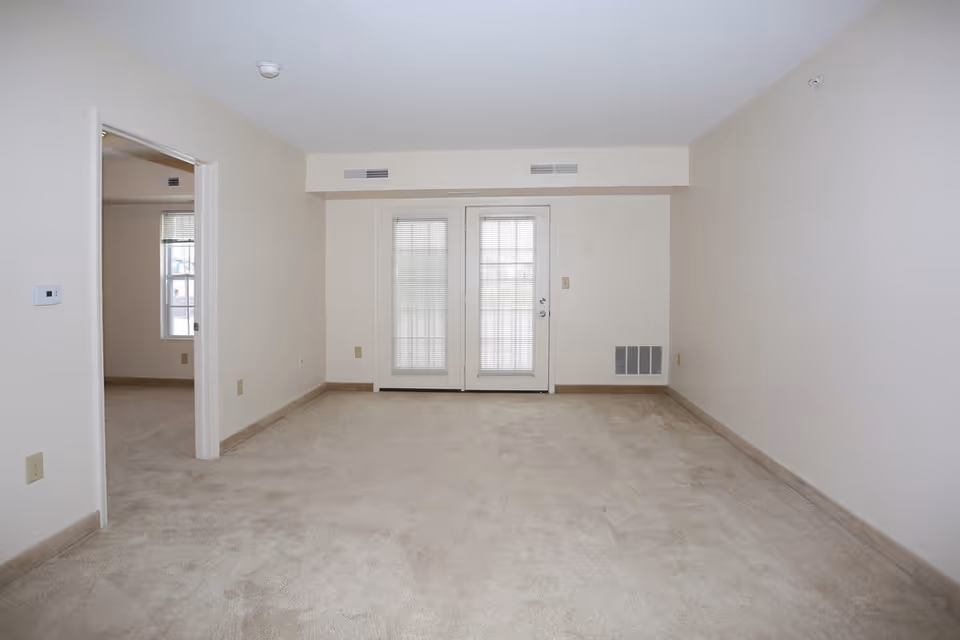 Empty room with beige carpet and off-white walls, featuring a set of double glass doors with blinds in the center wall. To the left, there is an open doorway leading to another room with a window. The room has a simple, clean appearance with ceiling vents and electrical outlets visible.