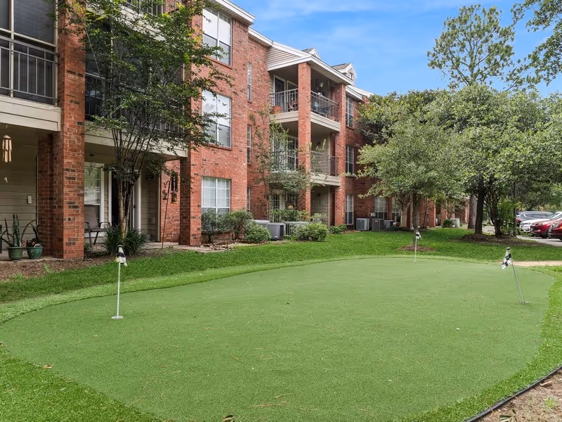 Outdoor putting green with three small flags in front of a red brick multi-story residential building with balconies and windows, surrounded by trees and parked cars.