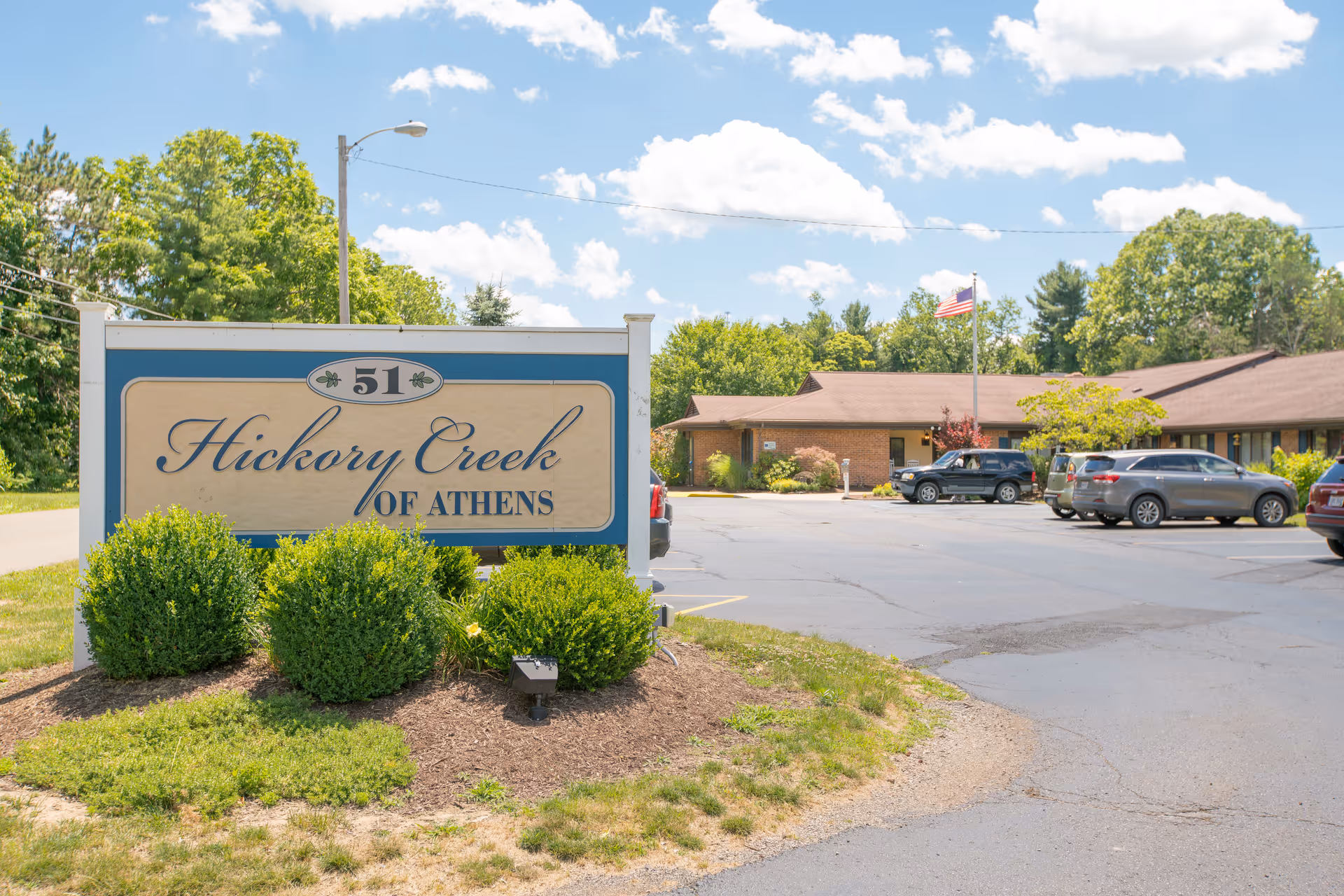 Outdoor view of Hickory Creek of Athens nursing center sign with bushes in front and a parking lot with several cars and a building with a flagpole in the background under a partly cloudy sky.