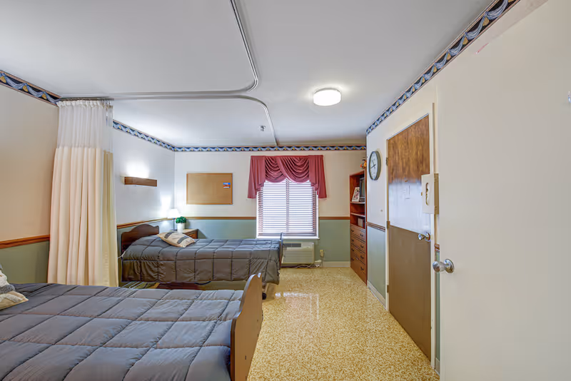 A senior living facility bedroom with two single beds covered in gray comforters. The room has a window with red curtains, a wooden door, a clock on the wall, a small bookshelf, and a curtain divider between the beds. The floor is covered with a speckled yellow and beige linoleum.