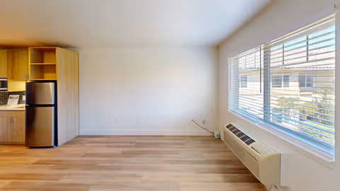 Empty studio-style living area with a small kitchenette and refrigerator on the left, wood floors, a large window with blinds and a wall-mounted air unit.
