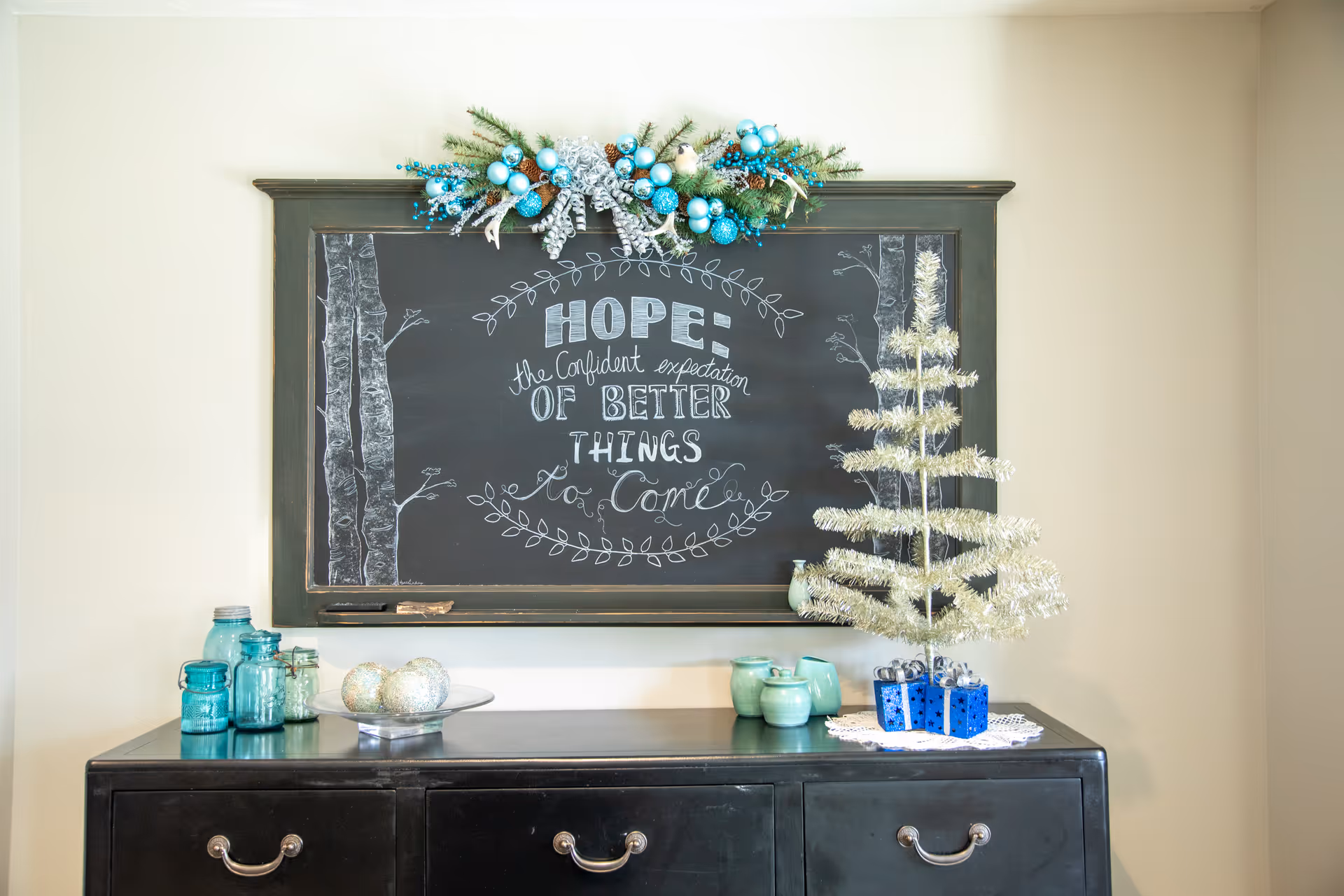 A black chalkboard with a decorative holiday garland on top and a handwritten inspirational quote that reads 'HOPE: the confident expectation of better things to come'. Below the chalkboard is a black dresser with blue and green glass jars, a plate with decorative silver balls, a small silver tinsel Christmas tree, and three small blue gift boxes.