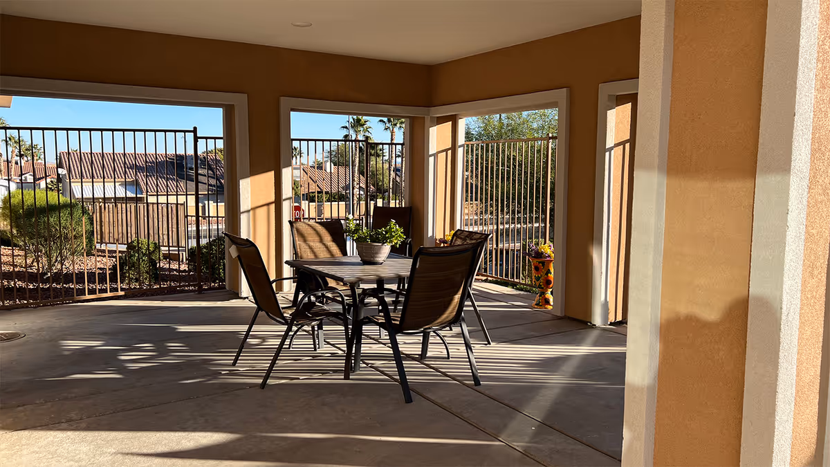 Covered outdoor patio area with a table and four chairs. The patio is enclosed by metal fencing and overlooks a landscaped area with bushes and trees. There is a potted plant on the table and a decorative flower pot near the fence.