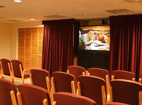 A small theater room with rows of wooden chairs upholstered in red fabric facing a large screen showing a movie scene with two people leaning over a table. The room has beige walls, a ceiling with recessed lighting, and red curtains framing the screen.