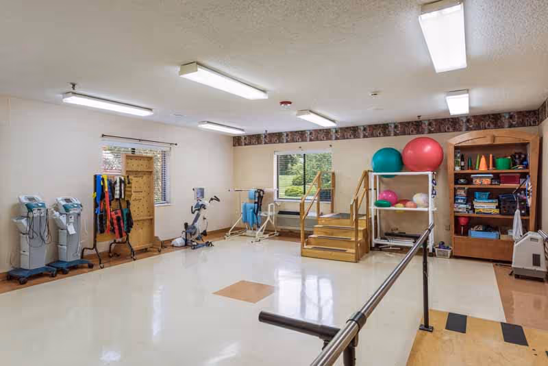 A rehabilitation therapy room with exercise equipment including stationary bikes, therapy bands, large exercise balls, and parallel bars. There are wooden stairs for physical therapy and a shelving unit with various therapy supplies. The room has windows letting in natural light and fluorescent ceiling lights.