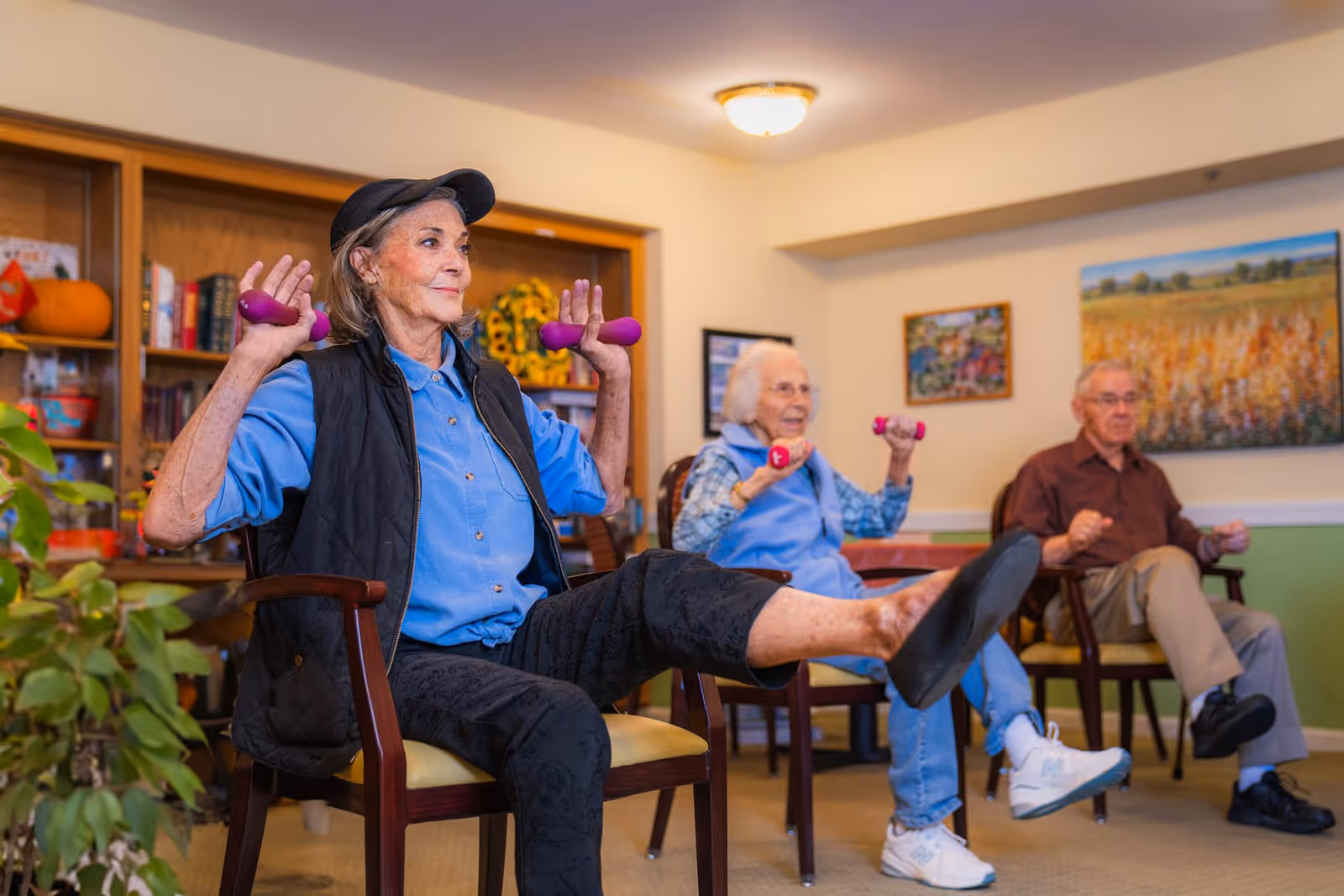 Three elderly individuals seated in chairs in a living room setting, participating in a seated exercise session using small purple hand weights. The woman in the foreground is lifting her leg and holding weights near her shoulders, while the two people in the background also hold weights and follow along. The room has bookshelves, framed artwork, and plants.