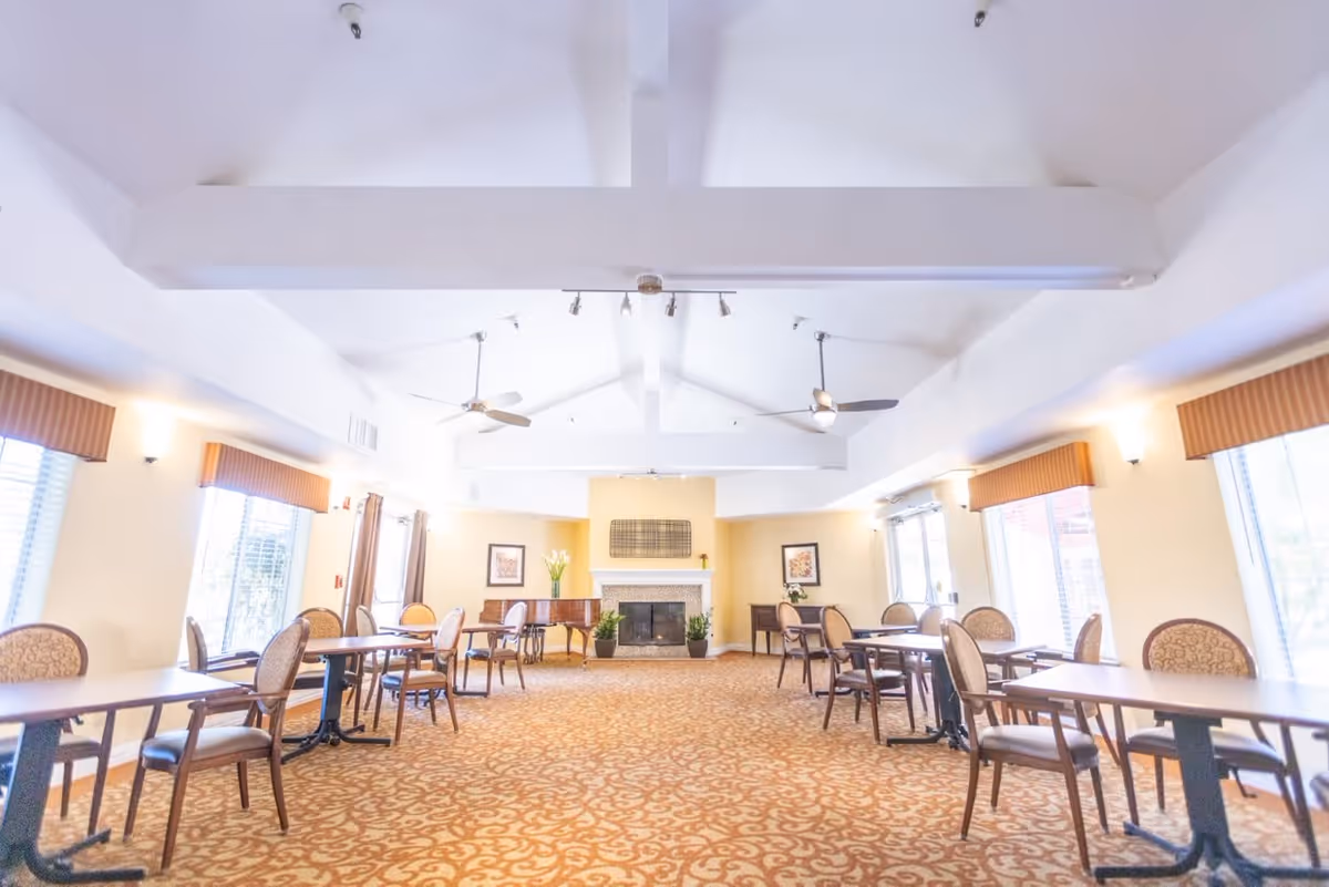A spacious, well-lit dining room with multiple tables and chairs arranged neatly. The room features large windows with valances, ceiling fans, a fireplace at the far end, and decorative framed artwork on the walls. The carpet has a patterned design in warm tones.