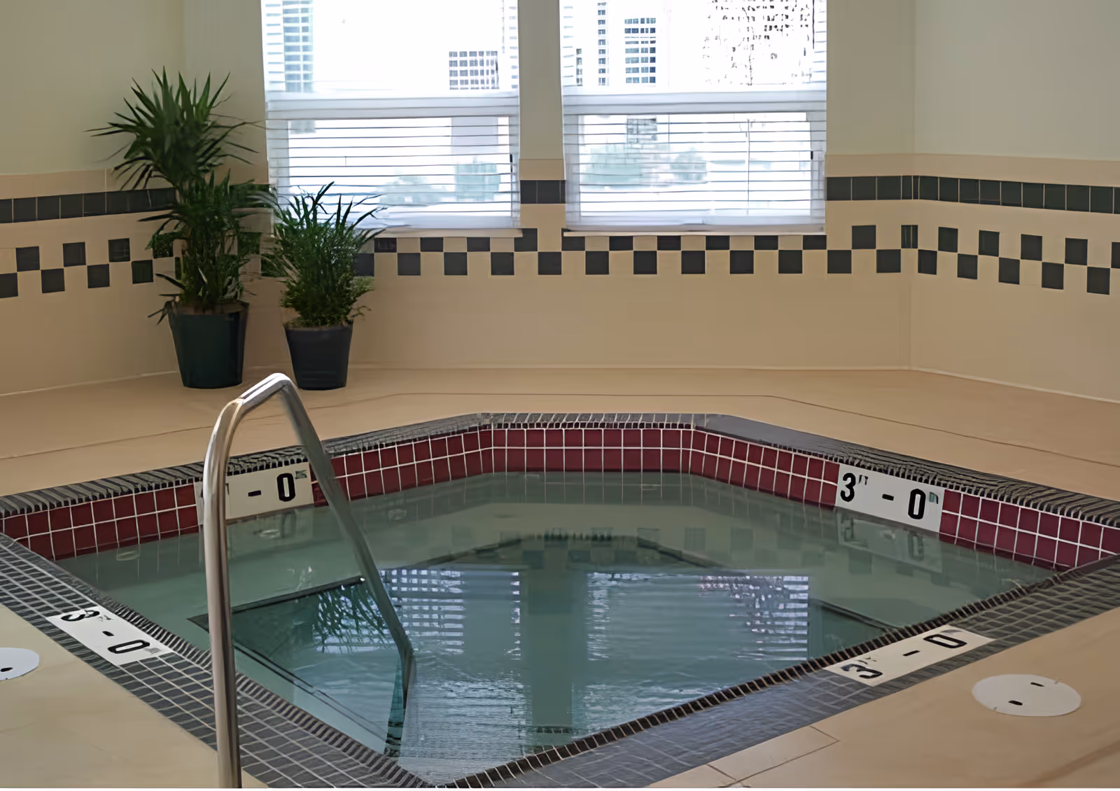 Indoor hot tub with tiled edges and metal handrail, surrounded by beige flooring and walls with a black and white checkered tile border. Two potted plants are placed near the windows with blinds.