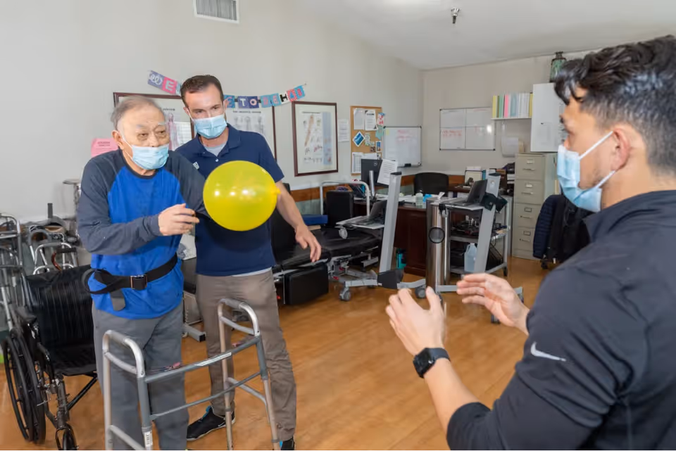 An elderly man using a walker and wearing a face mask is playing with a yellow balloon with the assistance of two caregivers, also wearing face masks, in a therapy or rehabilitation room with medical equipment and office furniture.