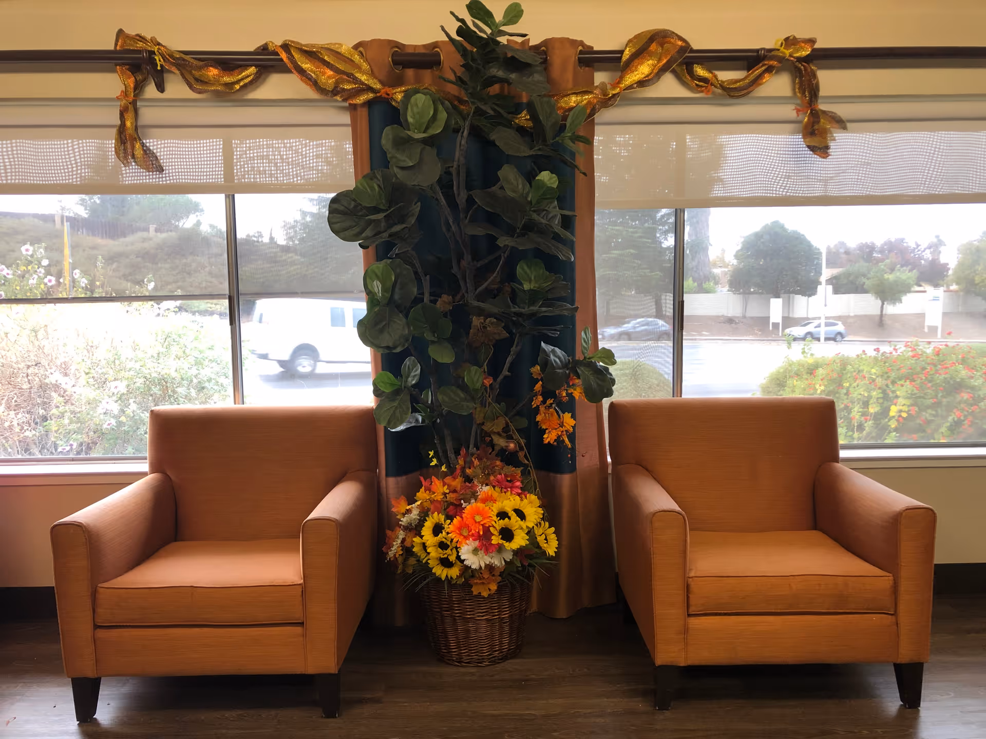 Two orange armchairs positioned side by side in front of large windows with a view of the outdoors. Between the chairs is a tall green plant and a basket of colorful flowers. The windows have beige curtains with a decorative gold and orange ribbon draped across the curtain rod.