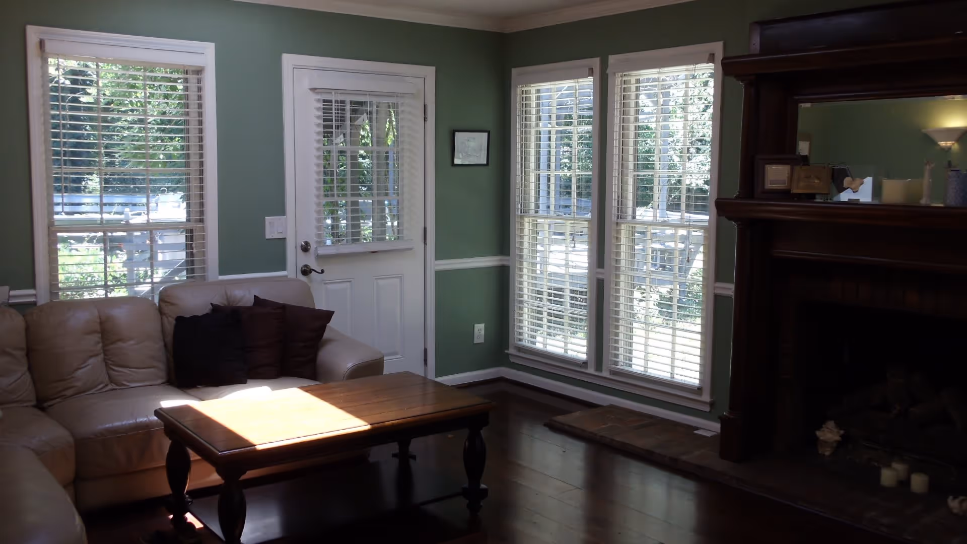 Sunlit living room with a beige sectional sofa, wooden coffee table, green walls, multiple windows and a fireplace.