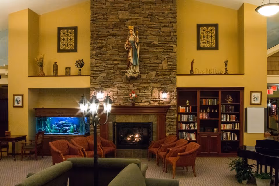 Cozy living room area with a stone fireplace in the center, a statue mounted above it, and a lit fire inside. Surrounding the fireplace are several orange armchairs and a green couch in the foreground. To the left, there is a large fish tank with aquatic plants and decorations. To the right, there is a wooden bookshelf filled with books and decorative items. The walls are painted yellow, and there are framed pictures and decorative pieces on shelves above the fireplace. A black lamp post style light fixture stands in front of the fireplace.