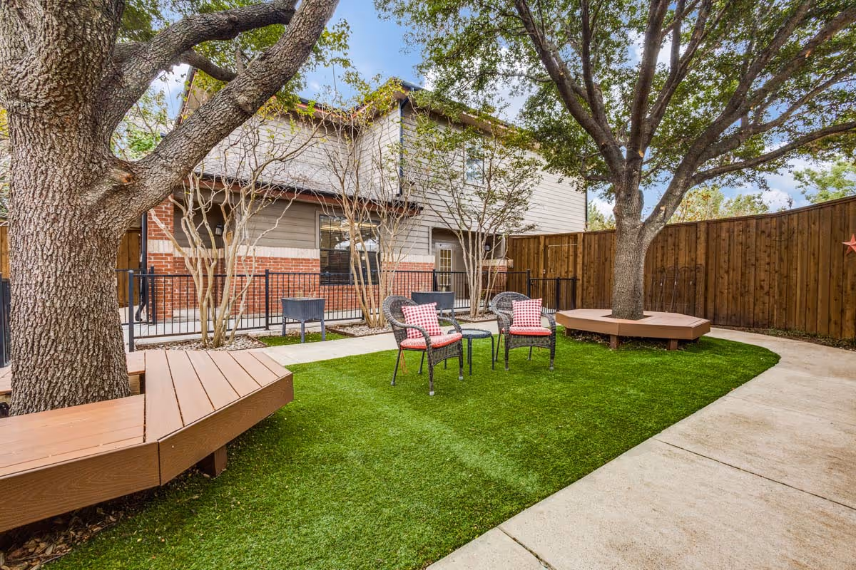 Outdoor seating area in a senior living facility with two large trees surrounded by wooden benches, two wicker chairs with red and white checkered cushions, a small round table, green artificial grass, a concrete pathway, and a wooden fence enclosing the space.