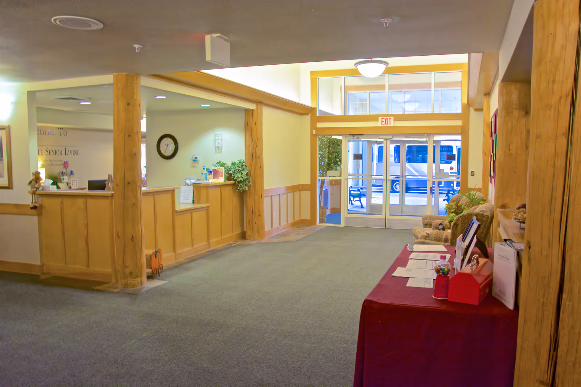 Interior view of a senior living facility lobby with a wooden reception desk on the left, a clock on the wall, and a glass exit door at the far end. There is a table covered with a red cloth on the right side with papers and a candy dispenser, and two upholstered chairs next to the table. The space is carpeted and has wooden beams and trim.