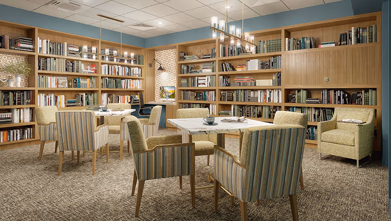 Well-lit communal library/reading room with wall-to-wall bookshelves, tables, and upholstered chairs.