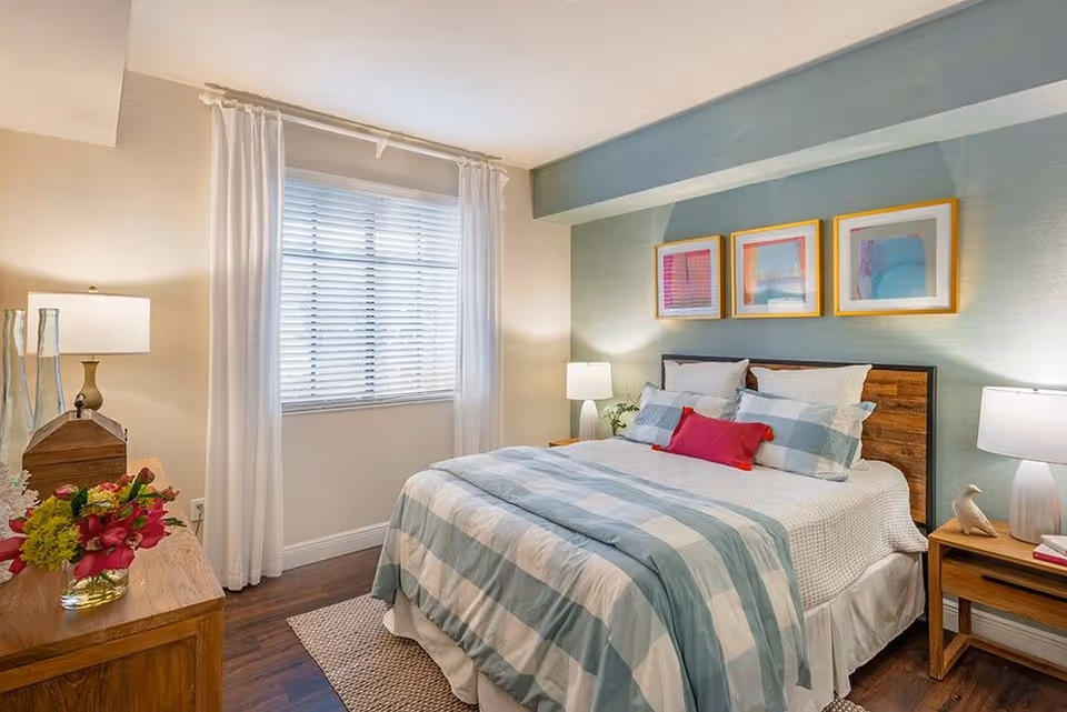 Well-lit bedroom with a bed dressed in blue-striped bedding, bedside tables and lamps, framed artwork above the headboard, a window with white curtains, and a wooden dresser with flowers.