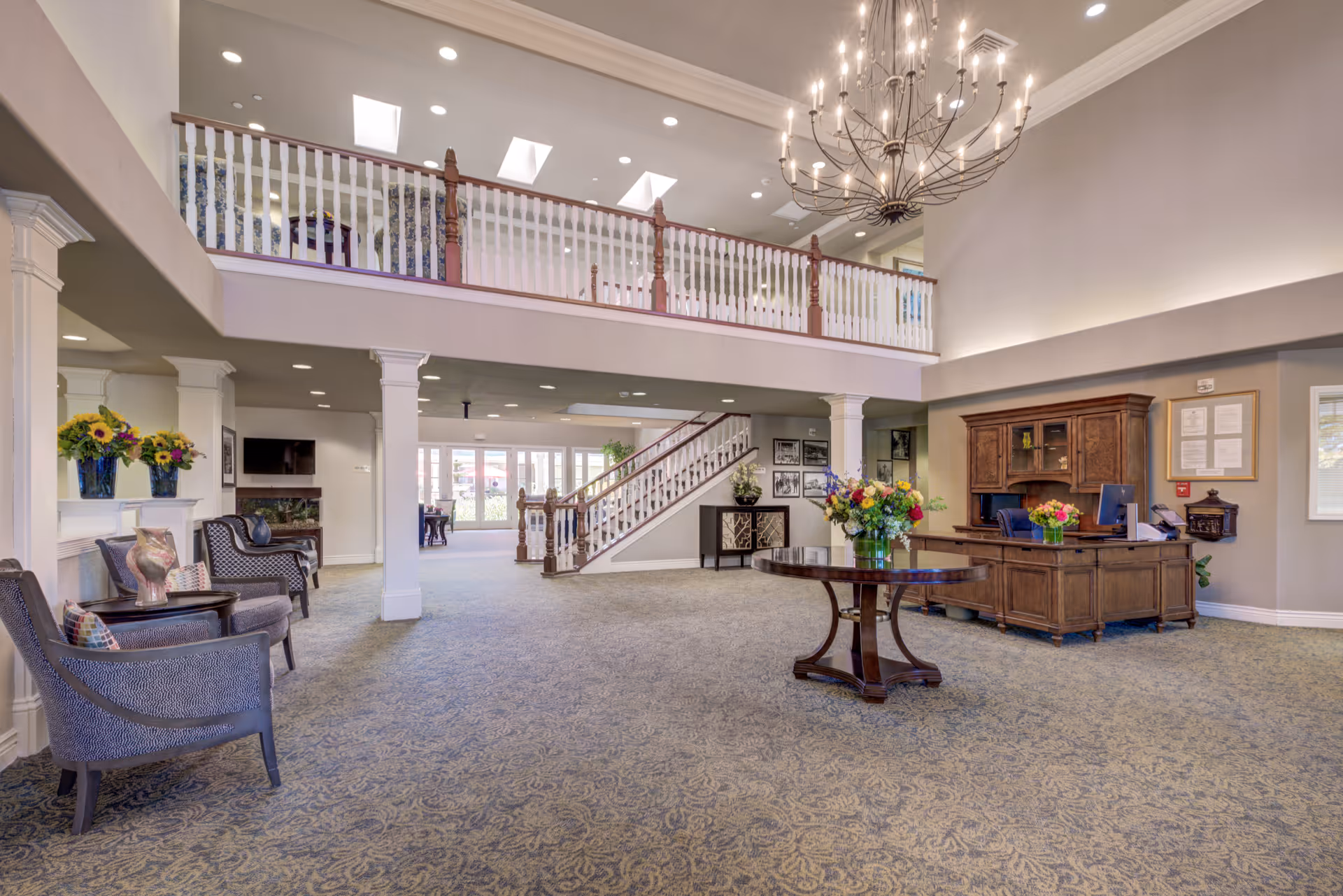 Spacious and elegant senior living facility lobby with high ceilings, a large chandelier, a round wooden table with a flower arrangement, a wooden reception desk, comfortable seating area with chairs and side tables, carpeted floor, and a staircase leading to an upper level with white railings.