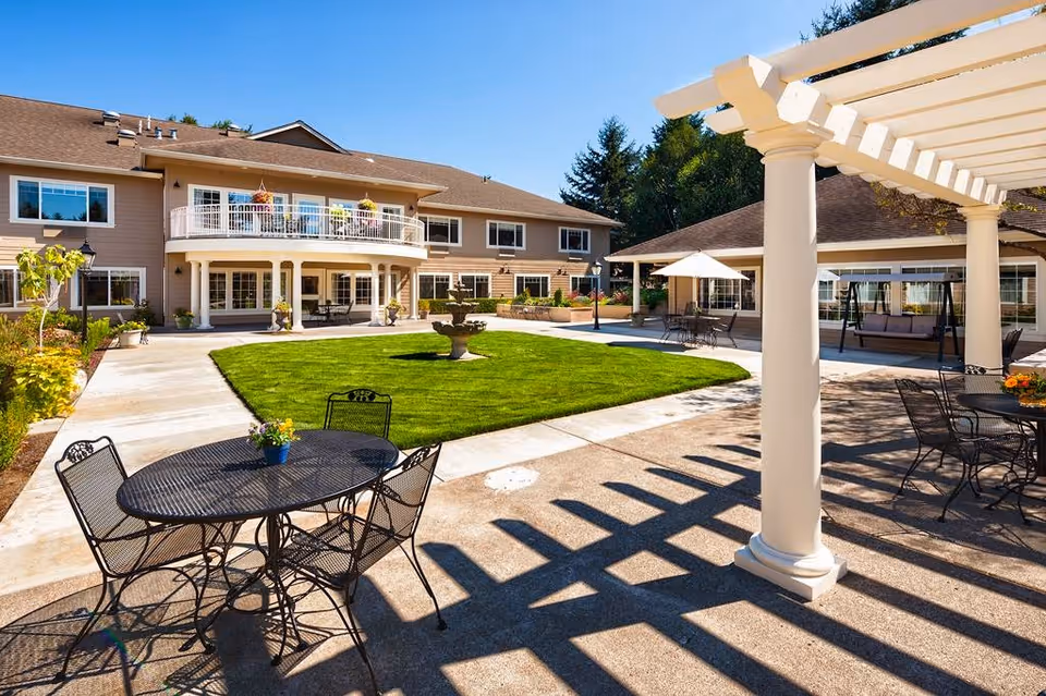 Outdoor courtyard area of a senior living facility with a green lawn, a central fountain, patio tables with chairs, a white pergola casting shadows, and a two-story building with balconies and large windows in the background under a clear blue sky.