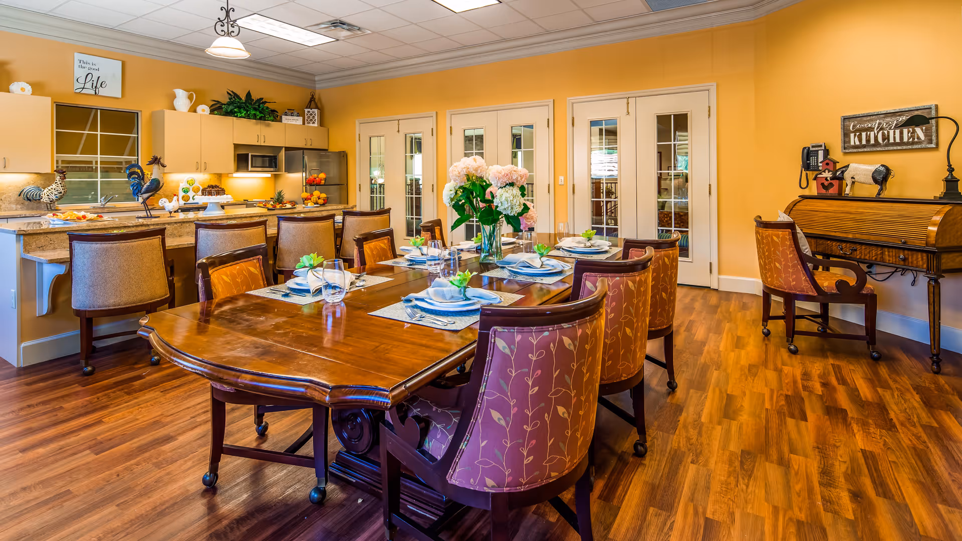 A warm and inviting dining area with a large wooden dining table set for six, featuring plates, glasses, and napkins with green decorative accents. The room has wooden flooring, yellow walls, and a kitchen area with a breakfast bar and stools in the background. There is a vintage roll-top desk with a chair on the right side and decorative items including flowers and rooster figurines on the kitchen counter.