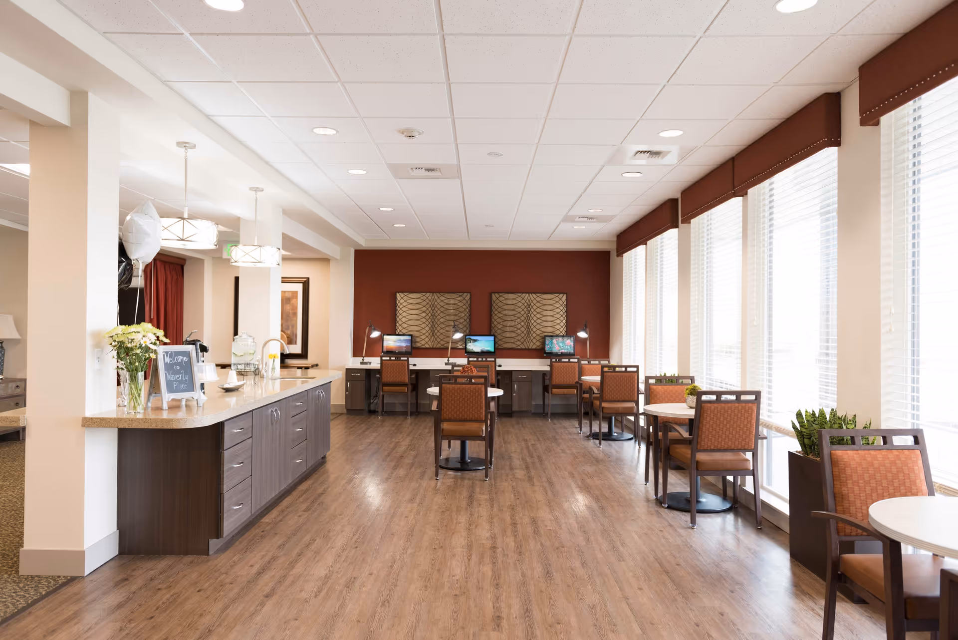 A bright and spacious common area in a senior living facility with large windows on the right side letting in natural light. The room features a long counter with a sink and flowers on the left, several small round tables with chairs along the windows, and a row of desks with computers and desk lamps against a red accent wall at the far end. The floor is wood, and the ceiling has recessed lighting.