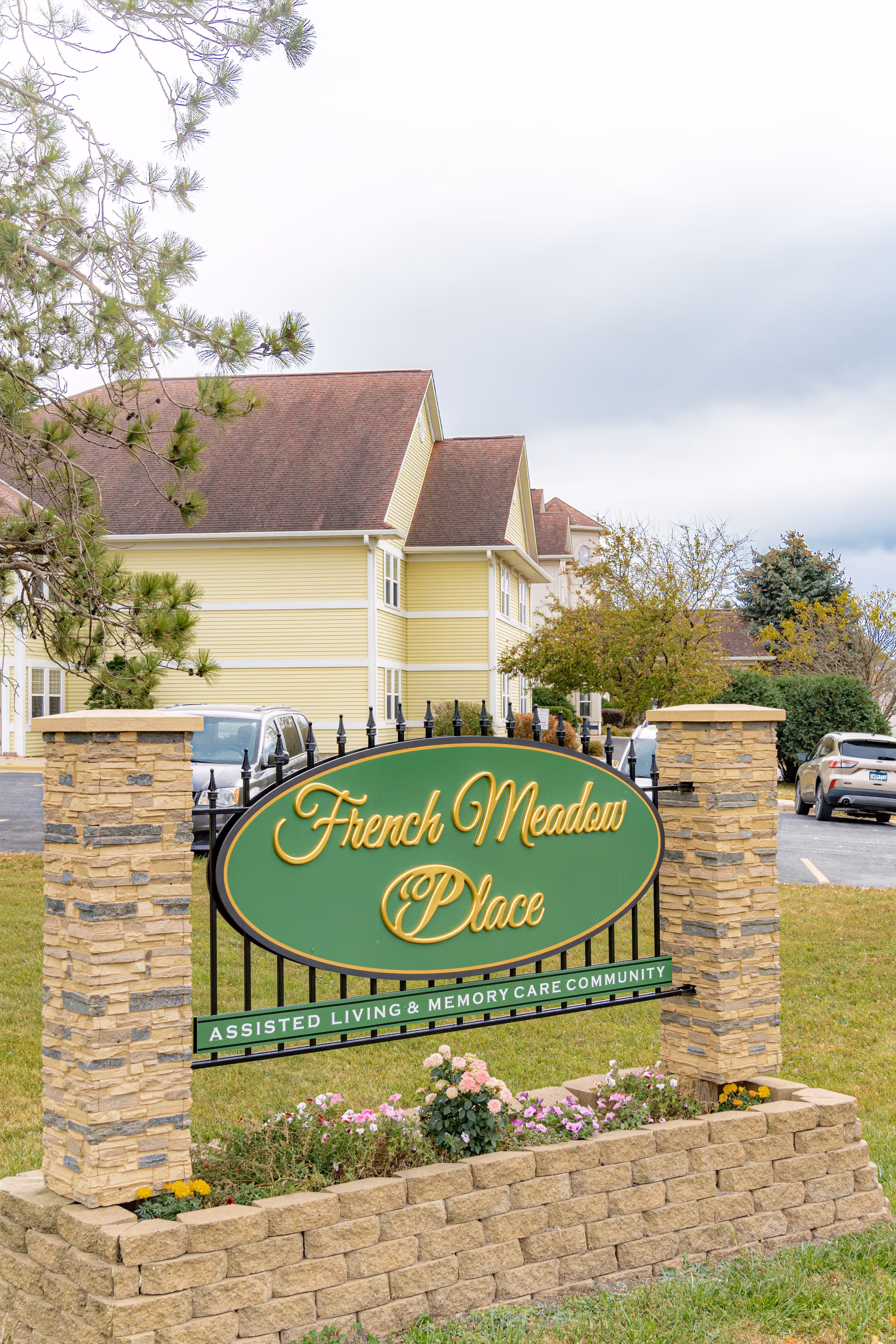 Outdoor view of the entrance sign for French Meadow Place, an assisted living and memory care community. The sign is green with yellow cursive text and is mounted between two stone pillars with a flower bed at the base. In the background, there is a yellow building with a brown roof and some parked cars.