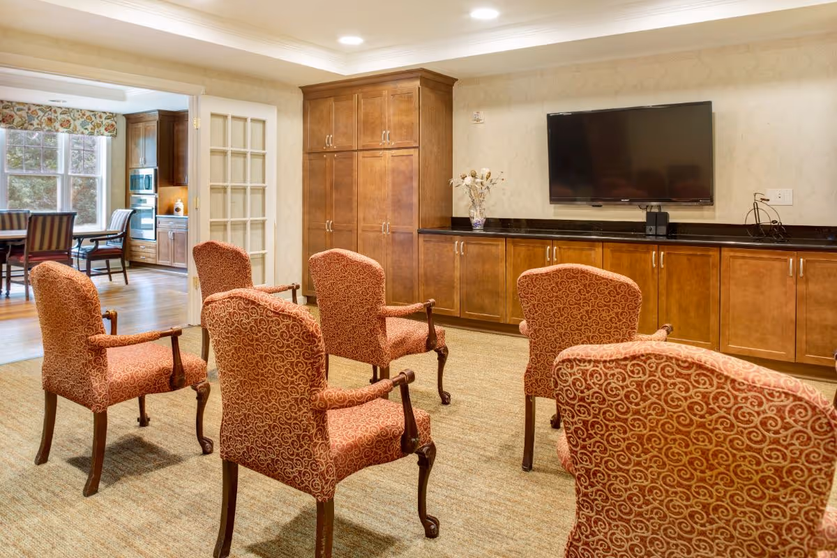A cozy common area with five upholstered armchairs arranged facing a wall-mounted flat screen TV. The room features wooden cabinetry along the wall beneath the TV and a beige carpet. Through an open doorway, a dining area with a table and chairs is visible, along with a kitchen area with wooden cabinets and appliances.