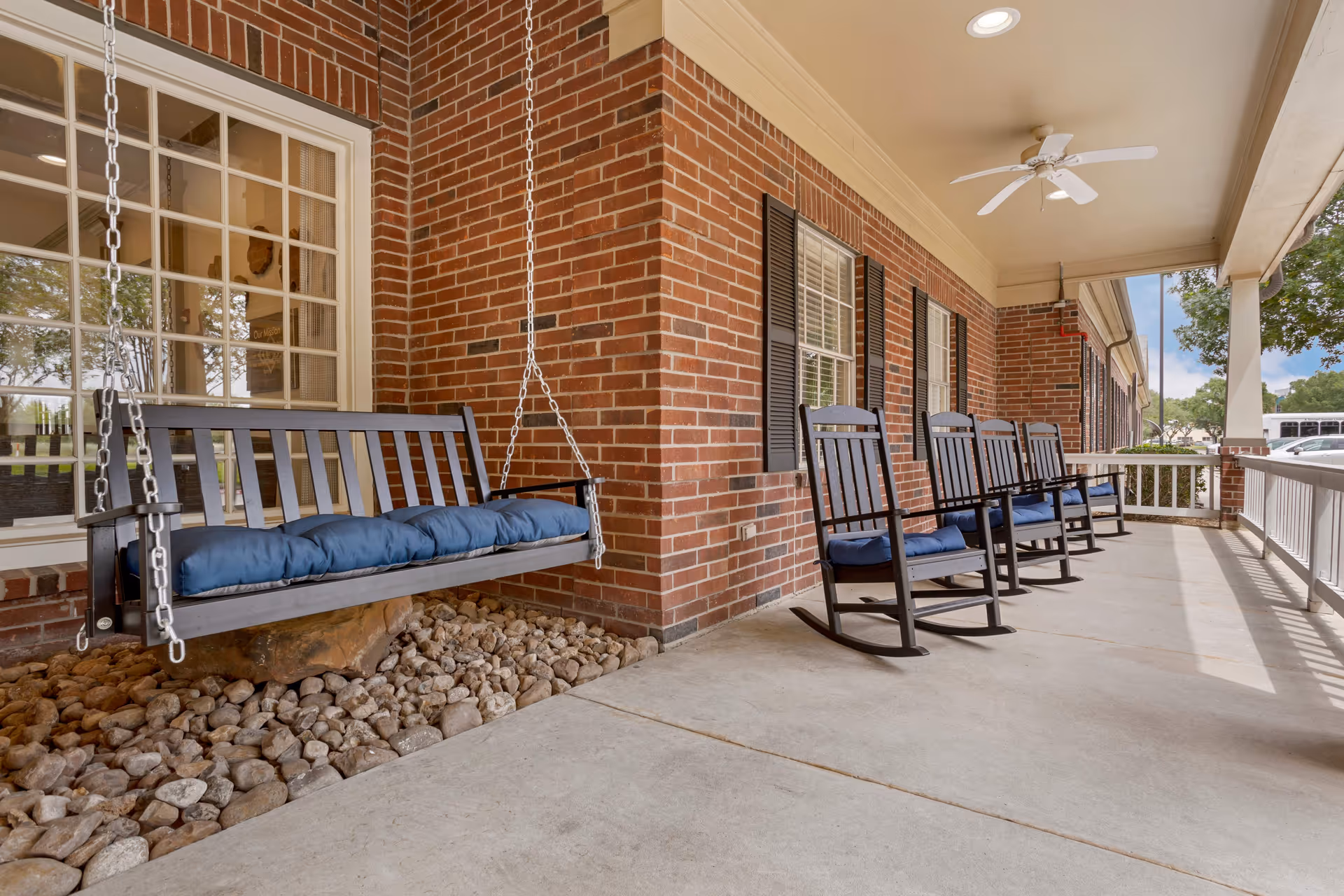 Covered brick front porch with a hanging bench swing and several rocking chairs with blue cushions.