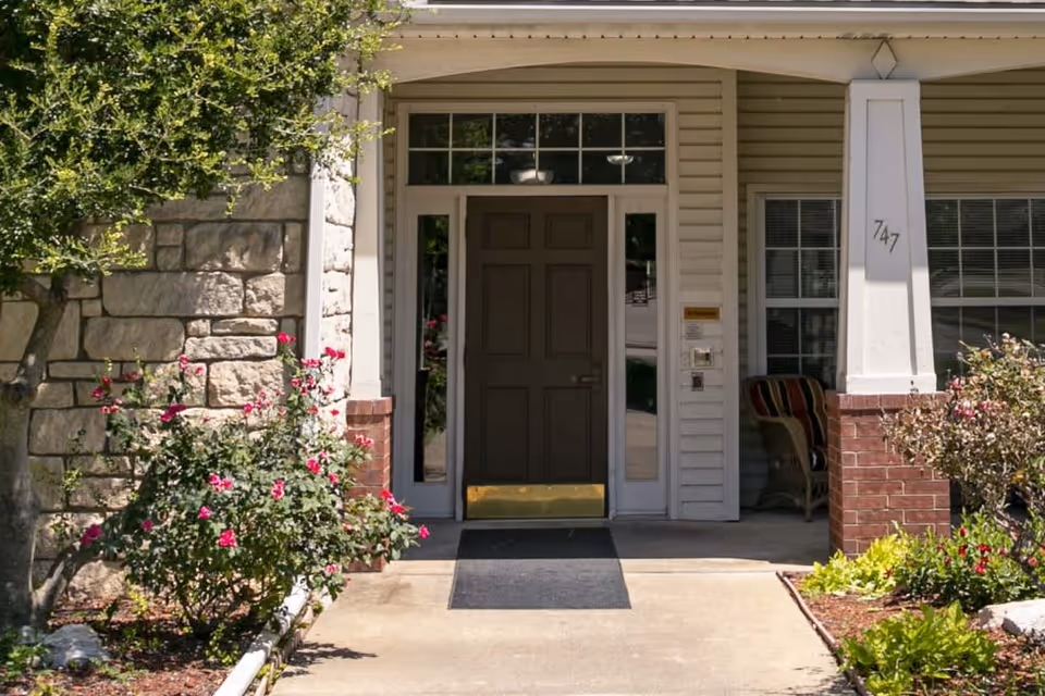 Entrance to a building with a dark brown door, sidelights, and a transom window above. The entrance is framed by a stone wall on the left and a white column with the number 747 on the right. There are flowering bushes and greenery on both sides of the concrete pathway leading to the door. A cushioned chair is visible on the porch to the right.