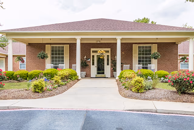 Front exterior view of a single-story brick building with a covered entrance supported by four white columns. There are two windows with white shutters on either side of the entrance, white rocking chairs on the porch, and well-maintained landscaping with bushes and flowers along the walkway leading to the door.