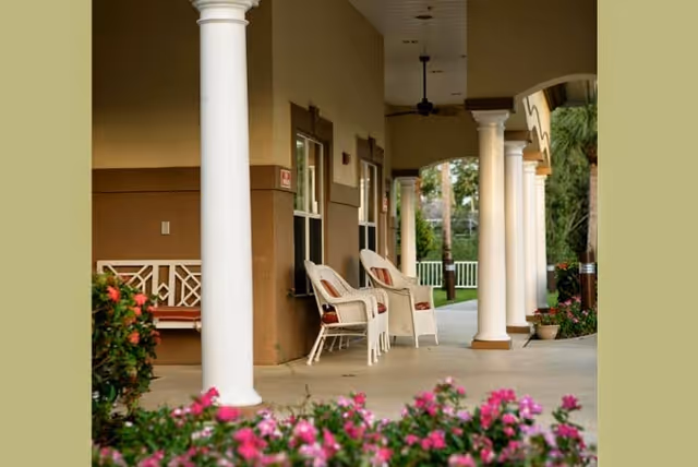 A covered outdoor patio area with white columns, two white wicker chairs with cushions, a bench, and flowering plants in the foreground. The patio overlooks a green lawn and trees in the background.