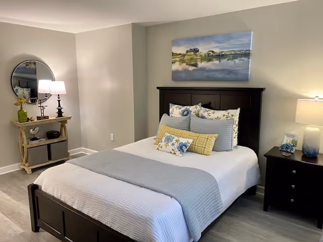 A neatly made bed with a dark wooden headboard and white bedding accented with blue and yellow pillows and a light blue throw blanket. To the right of the bed is a dark wooden nightstand with a lamp, a small picture frame, and a blue decorative item. On the left side of the room, there is a small wooden console table with a round mirror above it, a lamp, and some decorative items. The walls are painted light gray and the floor is a light wood laminate. A landscape painting hangs above the bed.