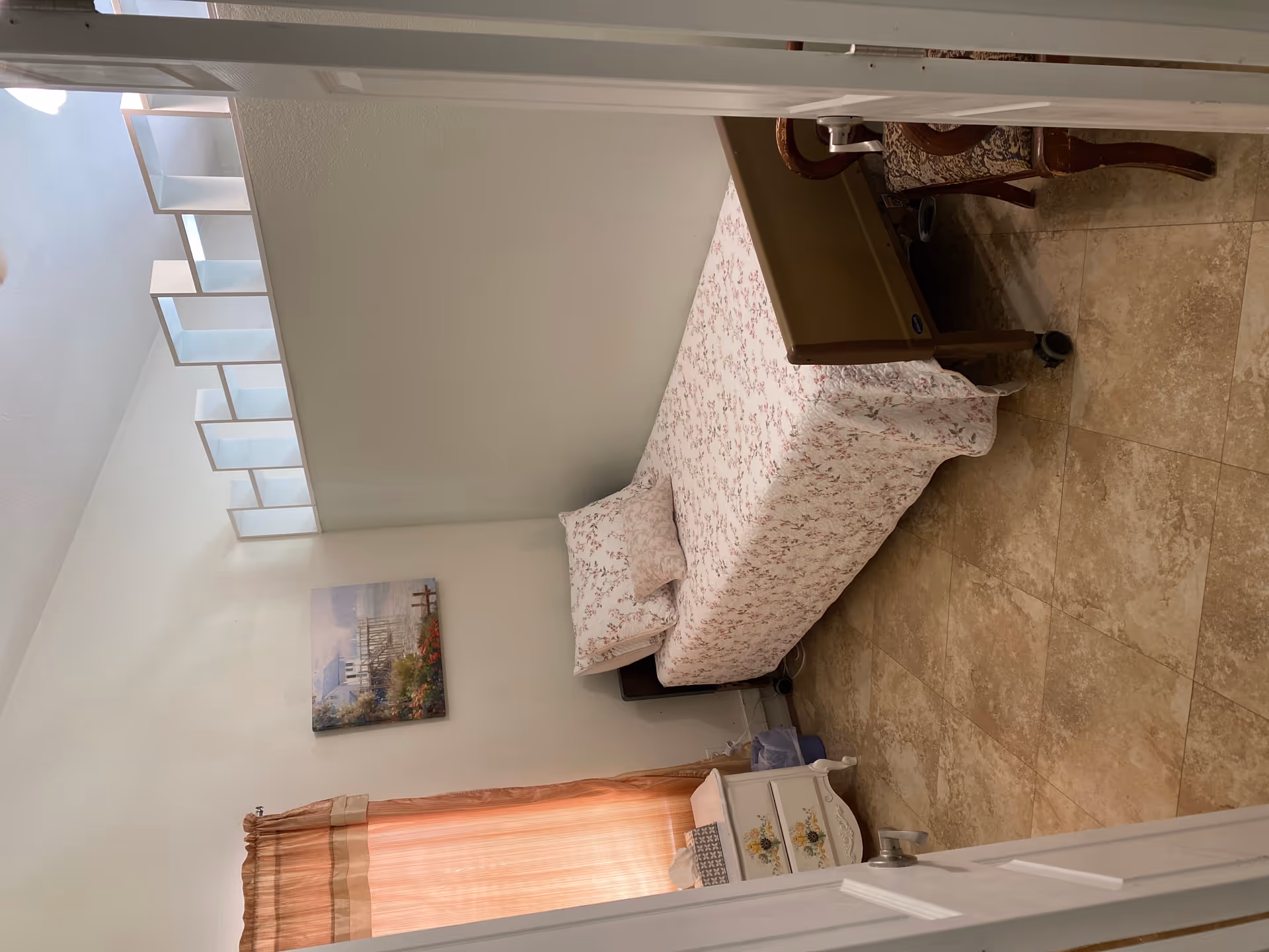 A small bedroom with a single bed covered in a floral bedspread and matching pillows. There is a white nightstand with floral designs on the drawers next to the bed, a window with peach-colored curtains, a painting of a lakeside house on the wall, and a wooden chair with patterned upholstery. The floor is tiled, and the room has a partial wall with open cubby-style shelving above it.