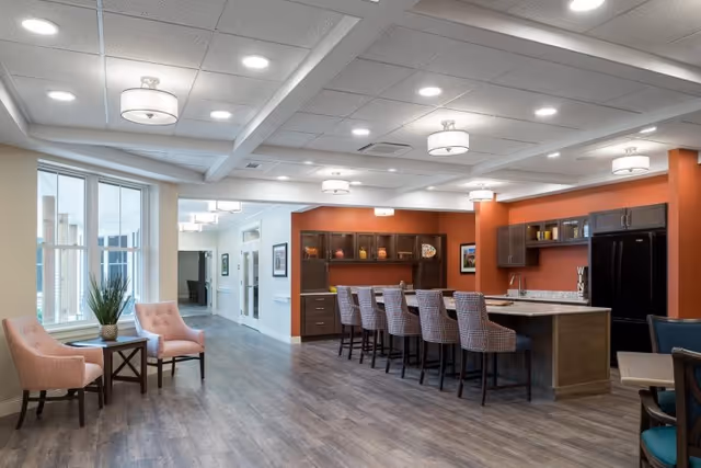 A bright communal dining and kitchen area with a long island and bar stools, seating by a window, and warm orange accent walls.