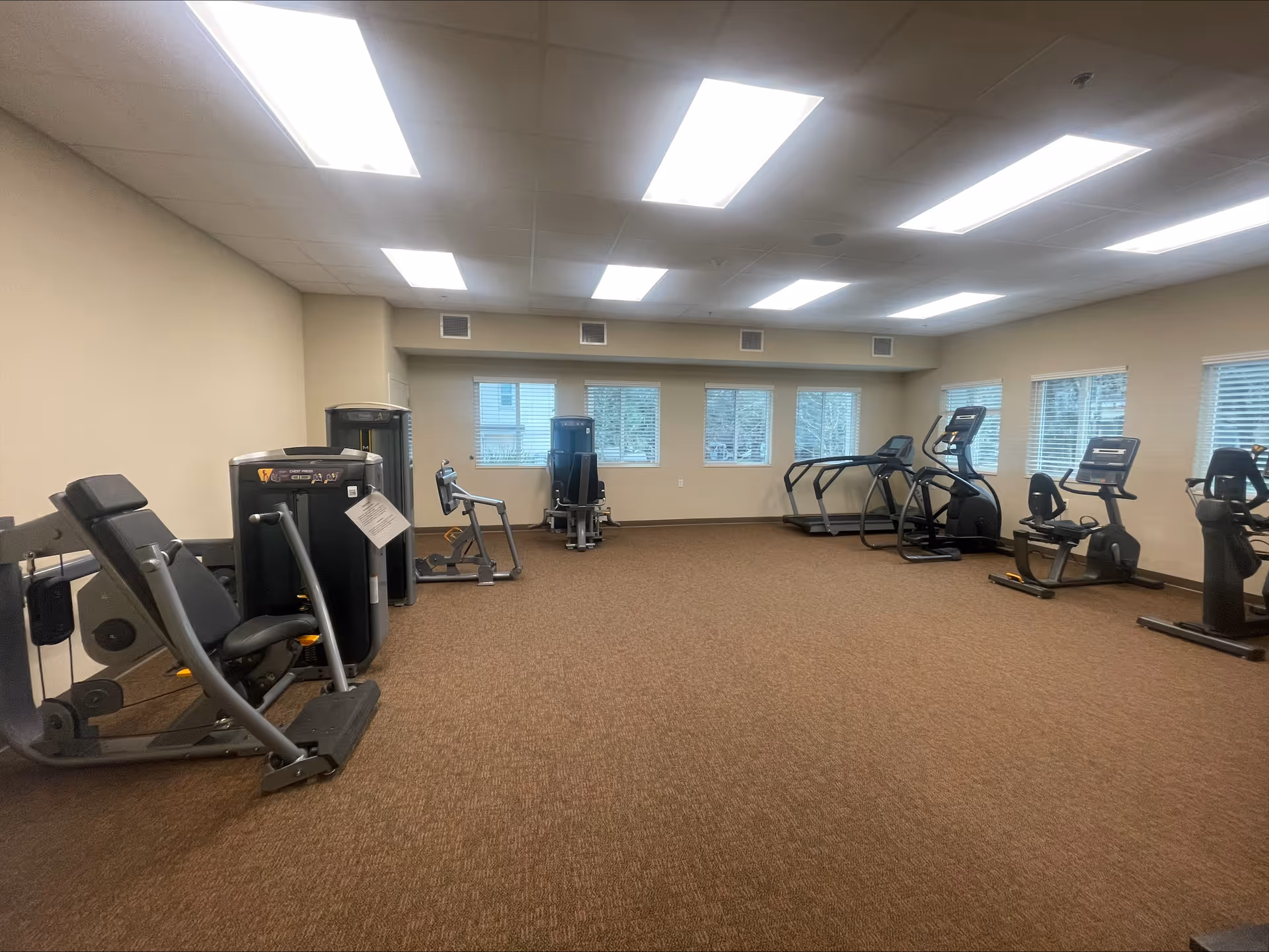 A spacious fitness room with various exercise equipment including weight machines, treadmills, elliptical machines, and stationary bikes. The room has beige walls, brown carpet flooring, and multiple windows with blinds allowing natural light to enter. The ceiling has several rectangular fluorescent lights.