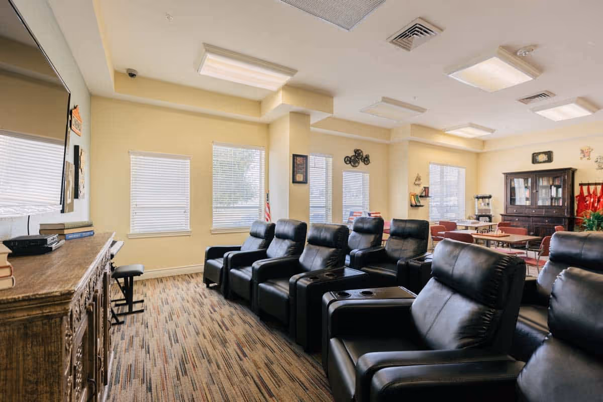 A bright and spacious common area in an assisted living facility featuring a row of black leather recliners with cup holders facing a large flat-screen TV mounted on the wall. The room has multiple windows with blinds, a wooden cabinet beneath the TV, a patterned carpet, and additional tables and chairs in the background for group activities or dining.