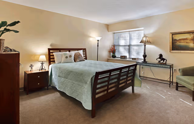 A neatly arranged bedroom with a wooden bed frame and a light green bedspread. There are two pillows on the bed, a nightstand with a lamp and a cross, a tall floor lamp, a table with decorative items including a horse statue, and a green armchair. A large window with blinds lets in natural light, and a framed landscape painting hangs on the wall.