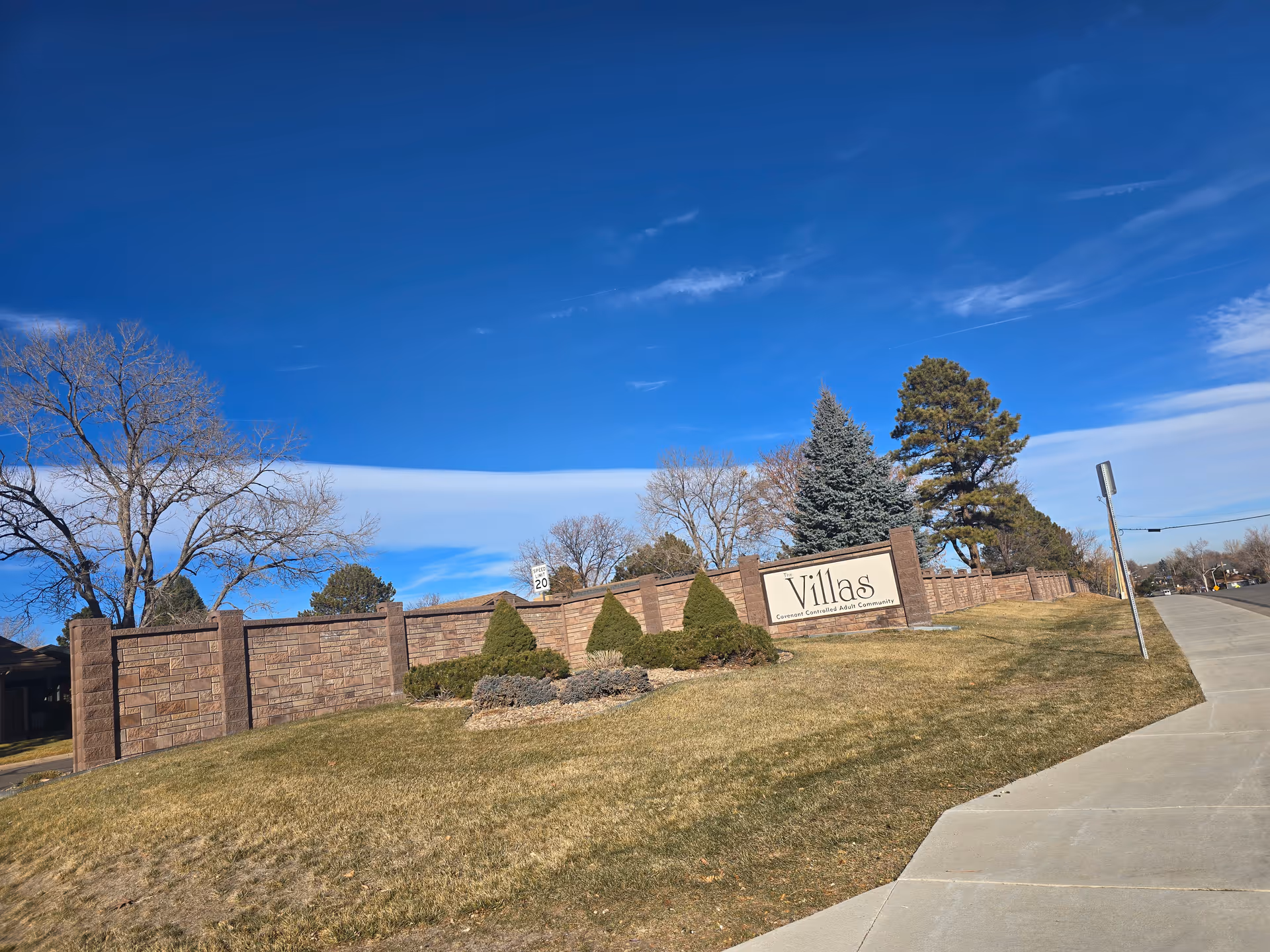 Brick entrance wall and sign reading "Villas" at the front of a senior living community with lawn, trees, and a blue sky.