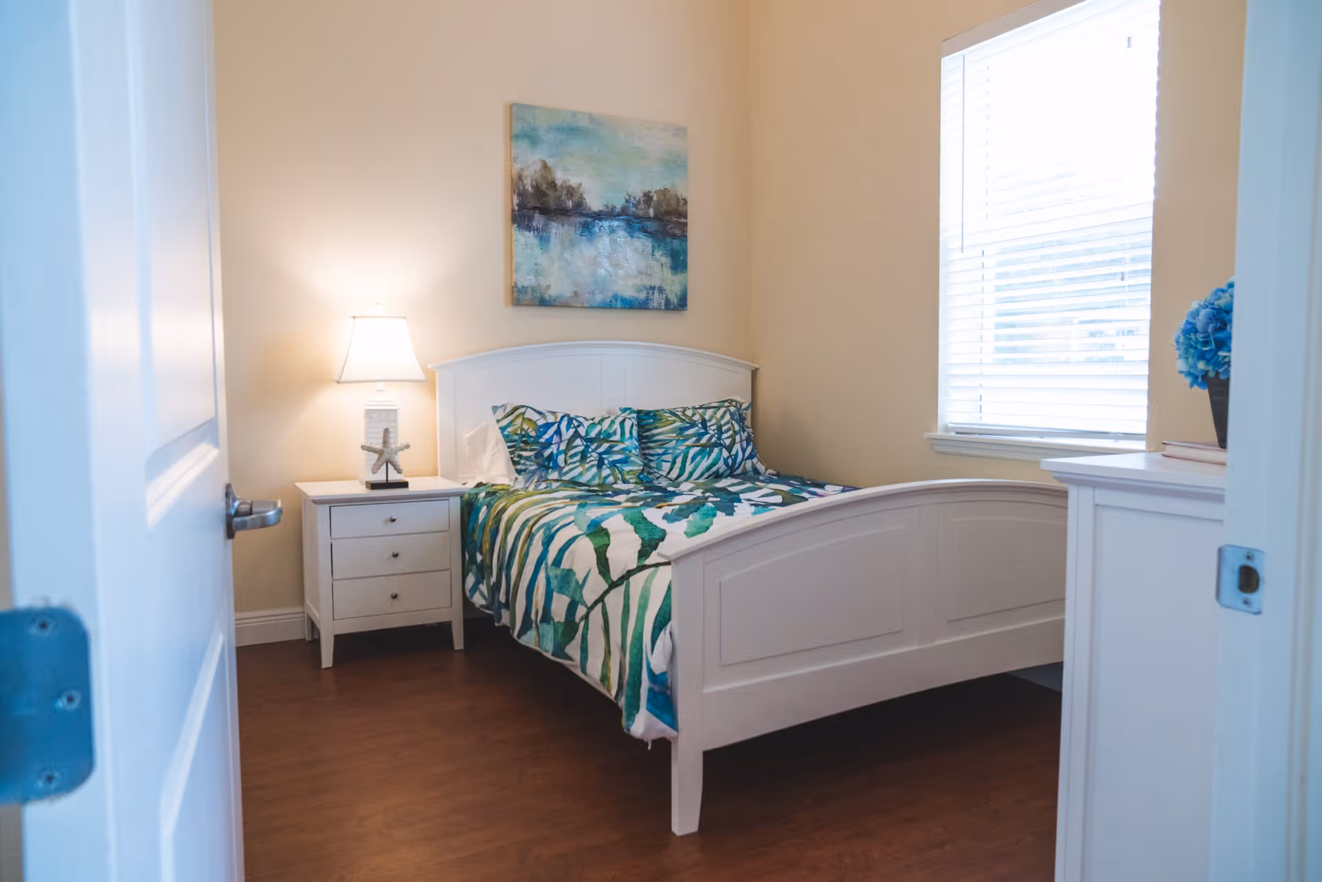 A bright bedroom with a white wooden bed frame and a bed made with green and blue leaf-patterned bedding. There is a white nightstand with a lamp and a starfish decoration on it next to the bed. A window with white blinds lets in natural light, and a piece of artwork depicting a serene landscape hangs on the wall above the bed.
