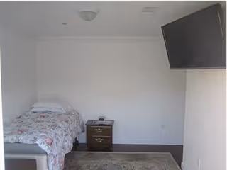 A small bedroom with a single bed covered in a floral bedspread, a wooden nightstand with two drawers and a clock on top, a wall-mounted flat-screen TV, and a patterned rug on the floor.