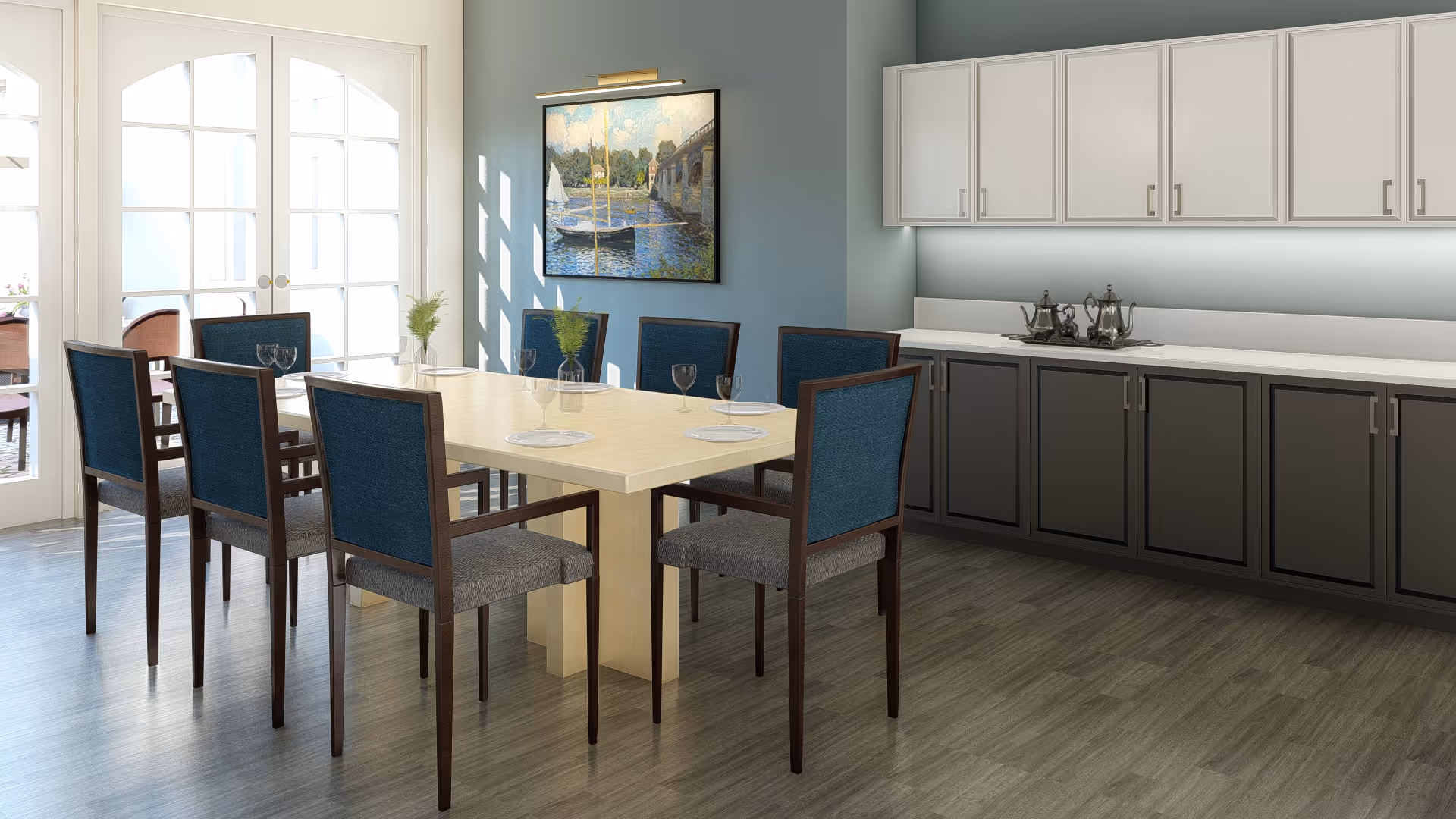 A dining room with a rectangular light wood table surrounded by eight chairs with blue backs and gray seats. The room has light blue walls, a large painting of a sailboat on the wall, white upper cabinets, and dark lower cabinets with a countertop holding a silver tea set. French doors lead to an outdoor patio area.