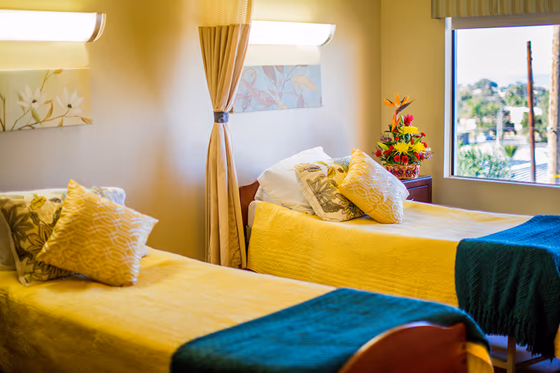 A bright and cozy room with two single beds covered in yellow bedspreads and green blankets at the foot. Each bed has multiple decorative pillows. A window on the right side lets in natural light and shows an outdoor view. A small table with a colorful flower arrangement is placed between the beds. The walls are decorated with floral artwork and a curtain is tied back between the beds.