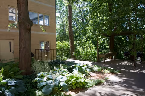 Outdoor garden area with lush green plants and trees next to a beige building. There is a paved pathway, a wooden bench, and two black charcoal grills under the shade of the trees.