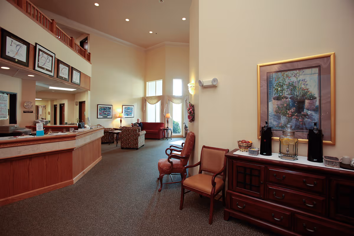 Interior view of a senior living facility lobby with a wooden reception desk on the left, seating area with armchairs and a sofa near large windows in the background, and a wooden sideboard on the right with beverage dispensers and a framed floral painting above it.