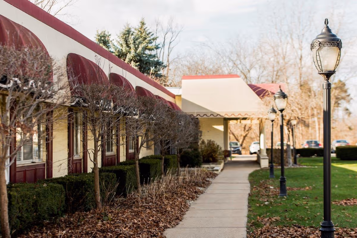 Sidewalk leading to the entrance of a building with maroon awnings over windows and a covered entryway. There are trimmed bushes and leafless trees along the building, and vintage-style street lamps line the path. Cars are parked in the background with a grassy area on the right side.