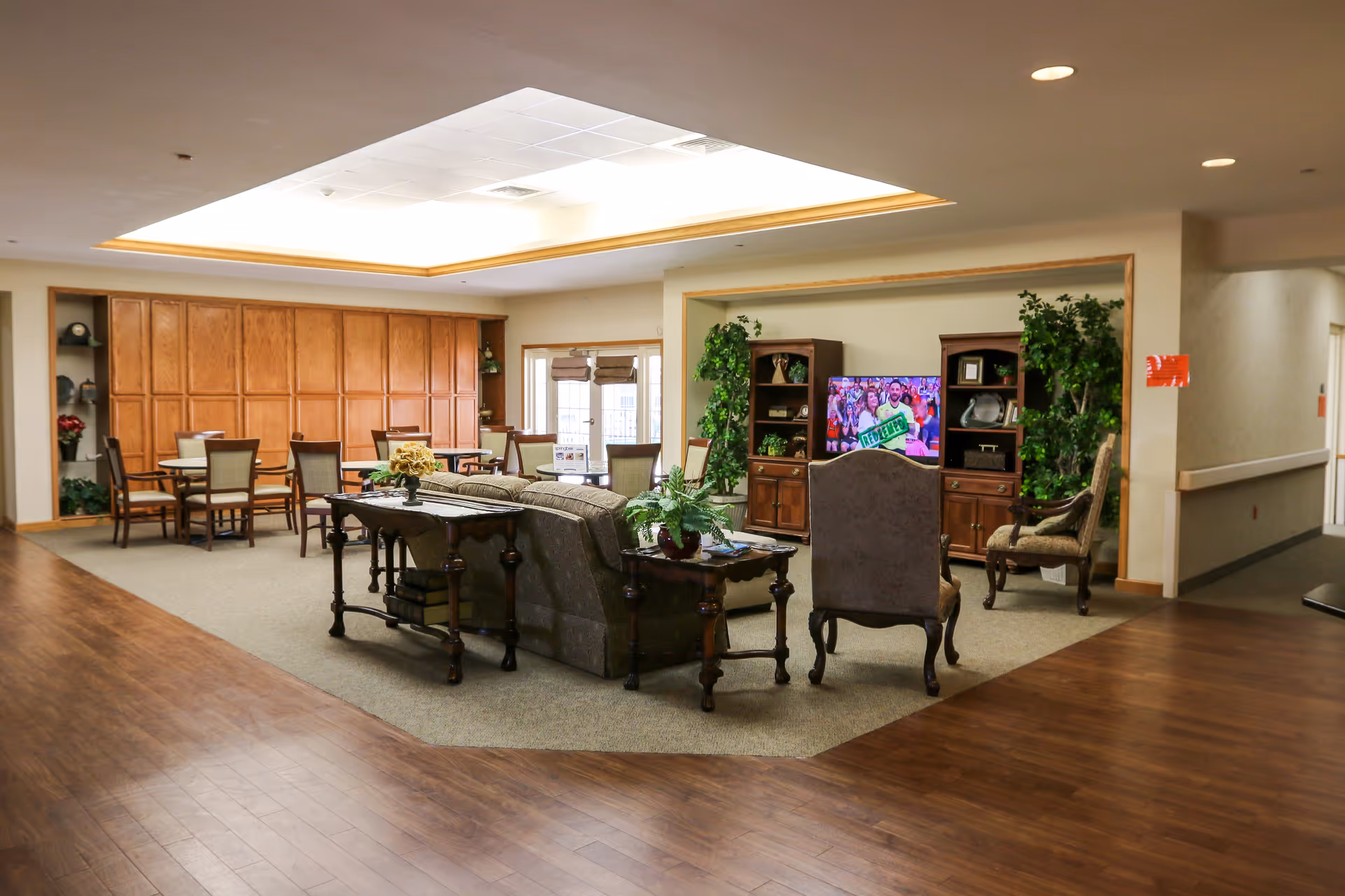 A spacious senior living common area with a seating arrangement including a sofa and chairs facing a television. The room features wood flooring, a carpeted seating area, wooden cabinets, dining tables with chairs, and decorative plants. The ceiling has a recessed lighting feature.